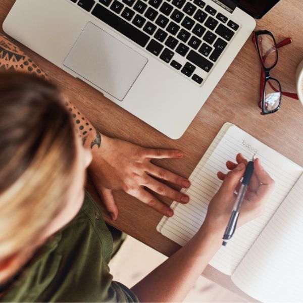 Woman writing in journal while sitting with laptop